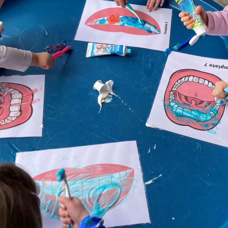 A blue table holds four laminated images of mouths with teeth. Children stand around the table cleaning the images with toothbrushes and toothpaste.